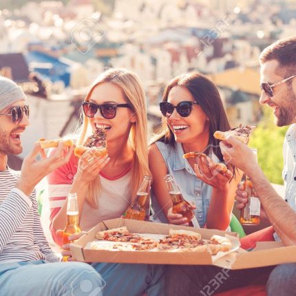 Pizza time. Four young happy people eating pizza and drinking beer while sitting at the bean bags on the roof of the building Pizza time. Four young happy people eating pizza and drinking beer while sitting at the bean bags on the roof of the building
